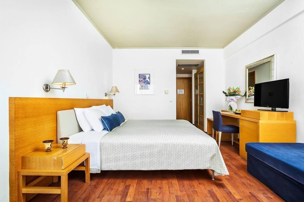 A hotel room with a bed, bedside tables, a desk with a chair, a television, and a small plant.
