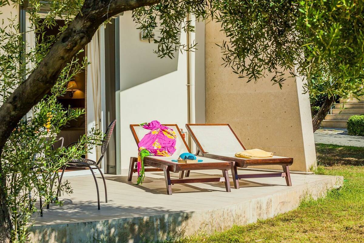 A sunlit outdoor seating area with two lounge chairs and a bench, surrounded by greenery and a building facade.