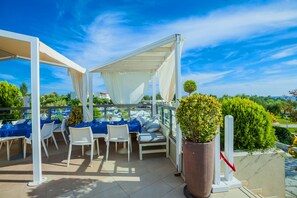 Une terrasse extérieure avec des tables et des chaises blanches, protégée par des auvents blancs, et agrémentée d’une plante en pot.