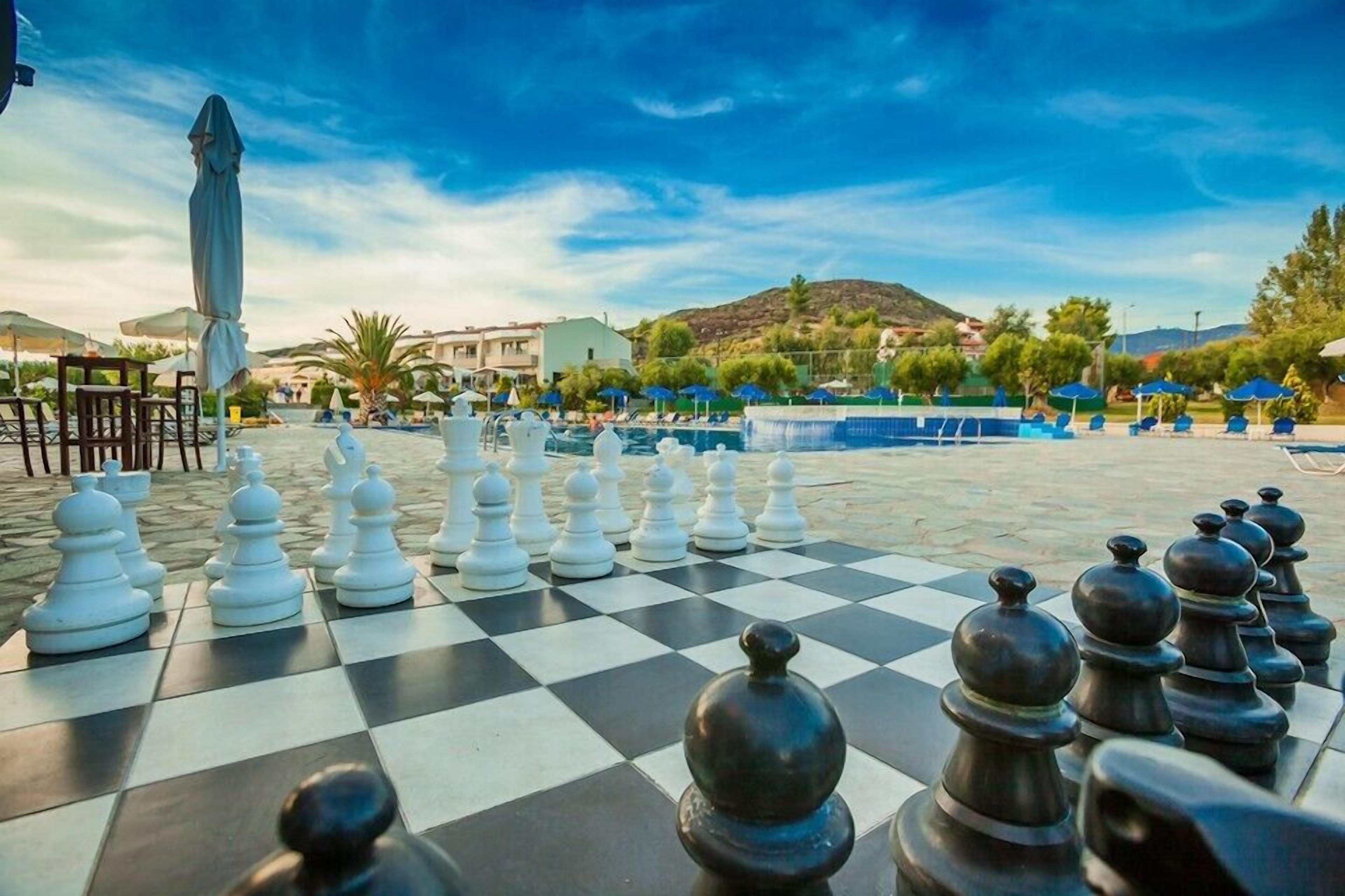 A chessboard set up outdoors with a pool and umbrellas in the background.
