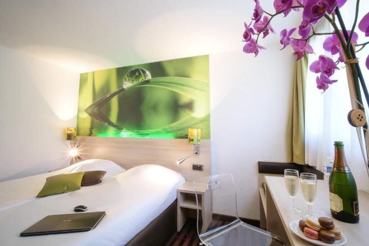 Une chambre d’hôtel avec un lit, un bureau sur lequel se trouvent une bouteille et des verres, un ordinateur portable et un tableau accroché au mur.