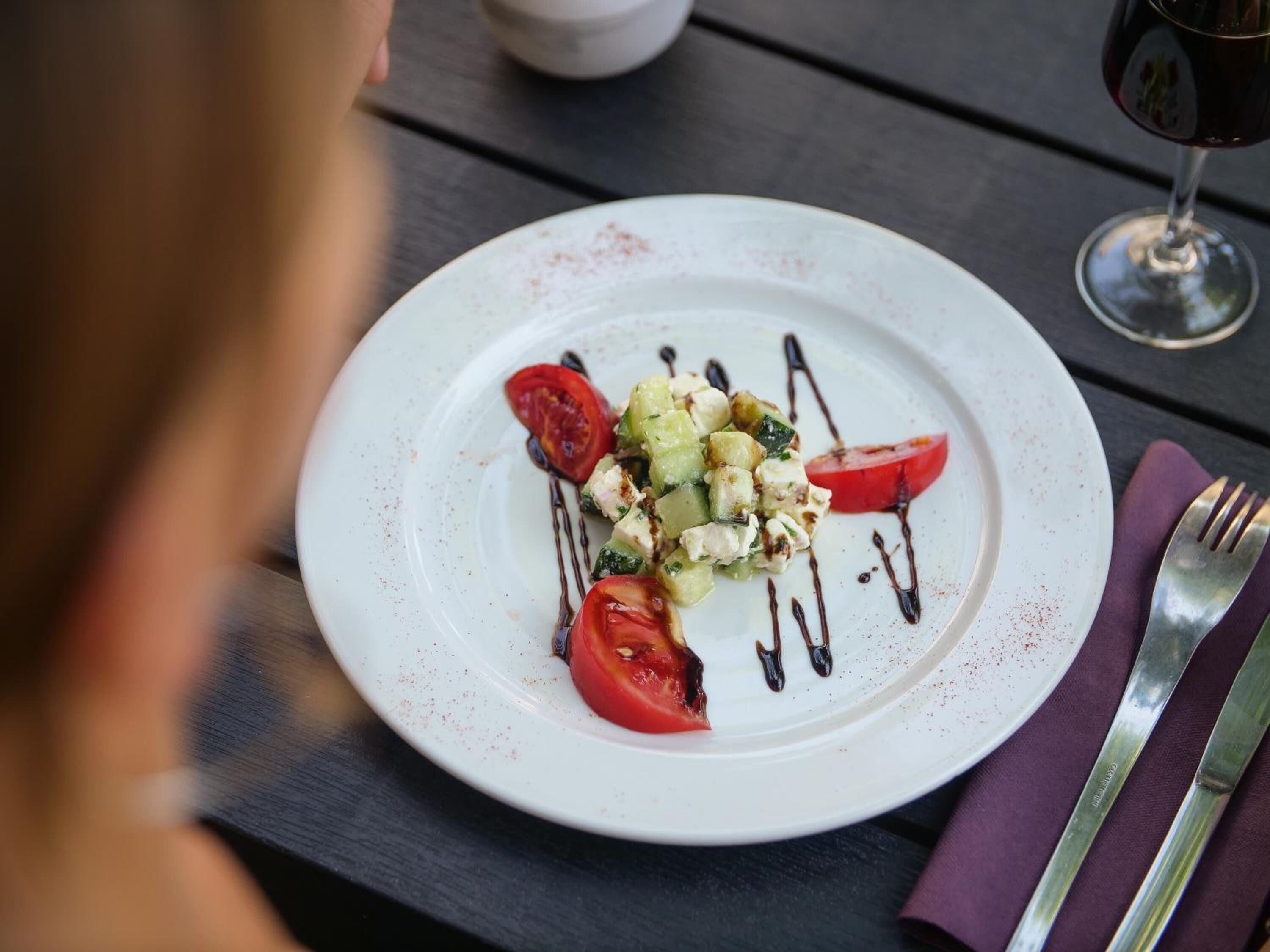 Un plato blanco con ensalada, una copa de vino tinto y cubiertos sobre una mesa de madera.