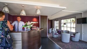 A hotel reception area with a curved desk, a receptionist, and a seating area with chairs and a TV screen.