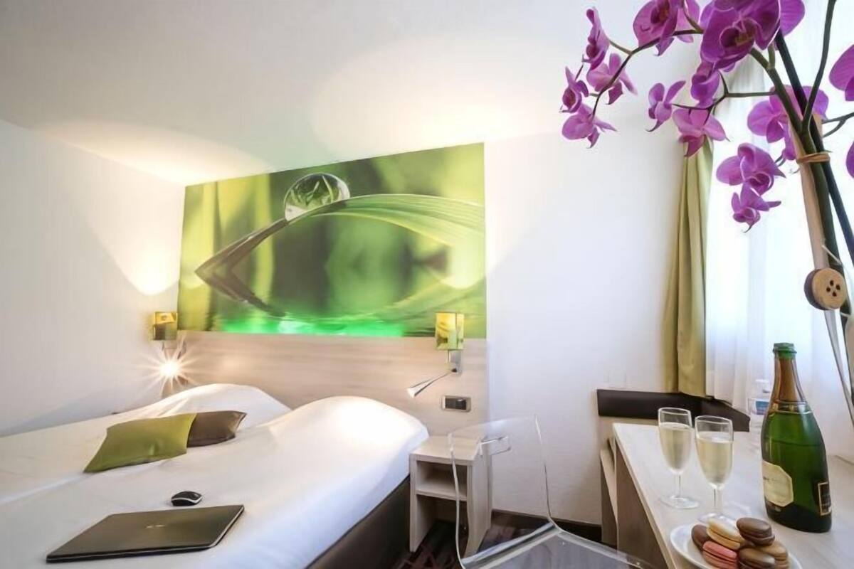 Une chambre d’hôtel avec un lit, un bureau avec un ordinateur portable, une bouteille de champagne et un vase de fleurs.