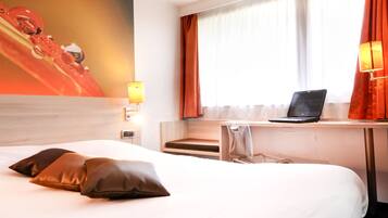 Habitación de hotel con cama, almohadas, escritorio con computadora portátil, lámpara y ventana con cortinas.