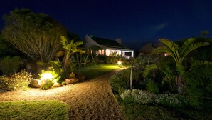 A nighttime view of a house with a gravel path leading to the entrance, surrounded by lush greenery and illuminated by garden lights. - Dune Ridge Country House (Saint Francis Bay)