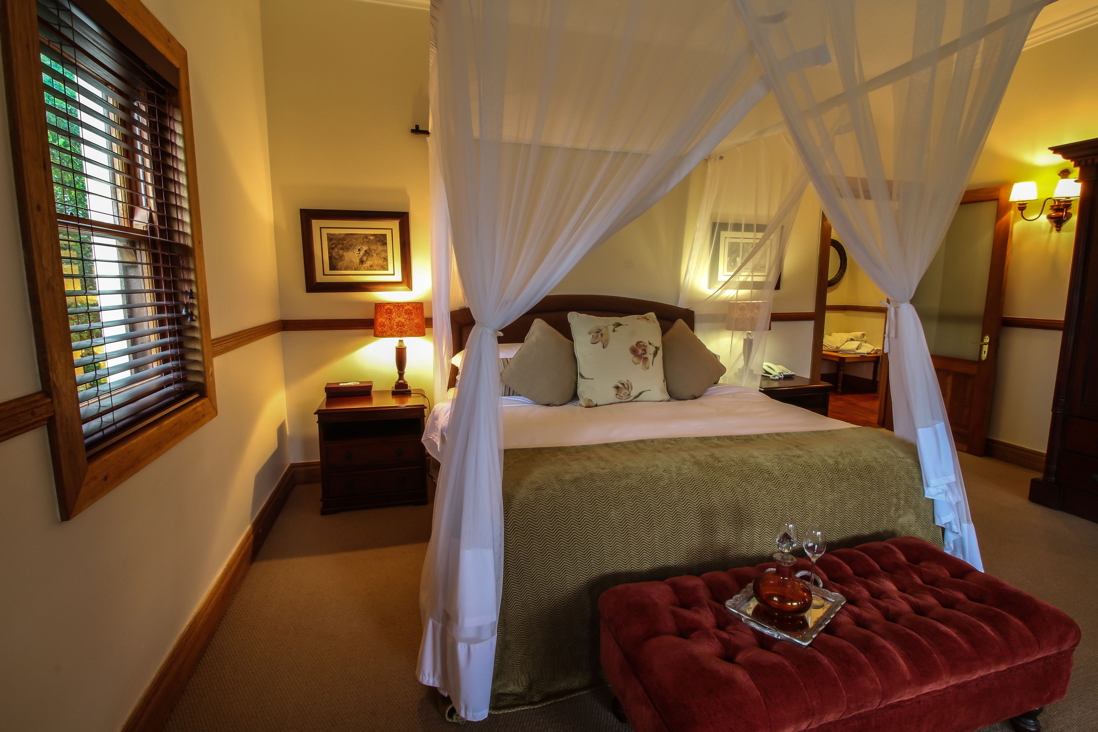 A bedroom with a four-poster bed, a nightstand with a lamp, and a red ottoman with a tray of wine glasses.