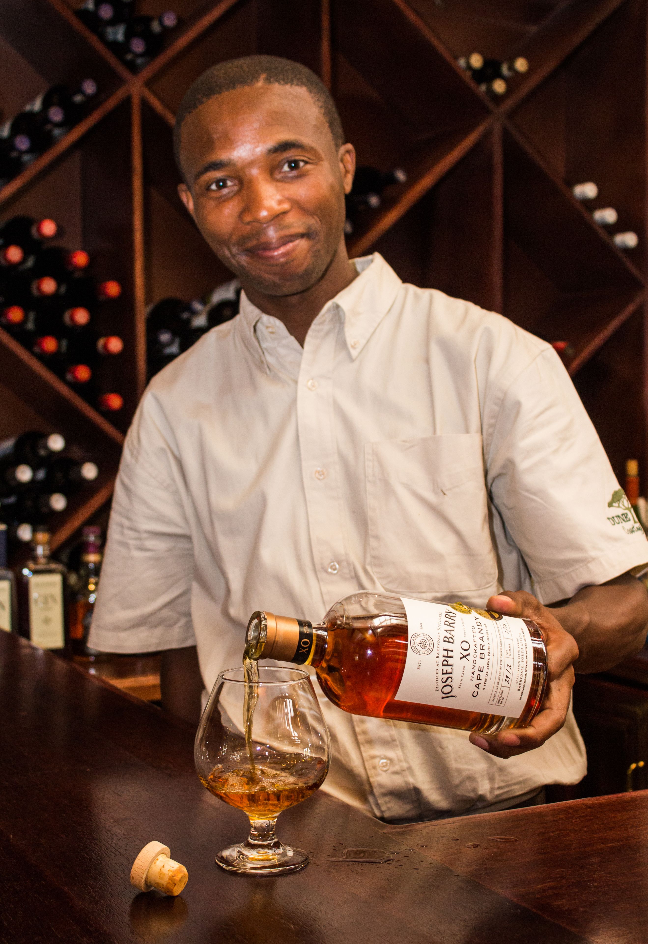 A person pouring a bottle into a glass at a bar with a wine rack in the background.