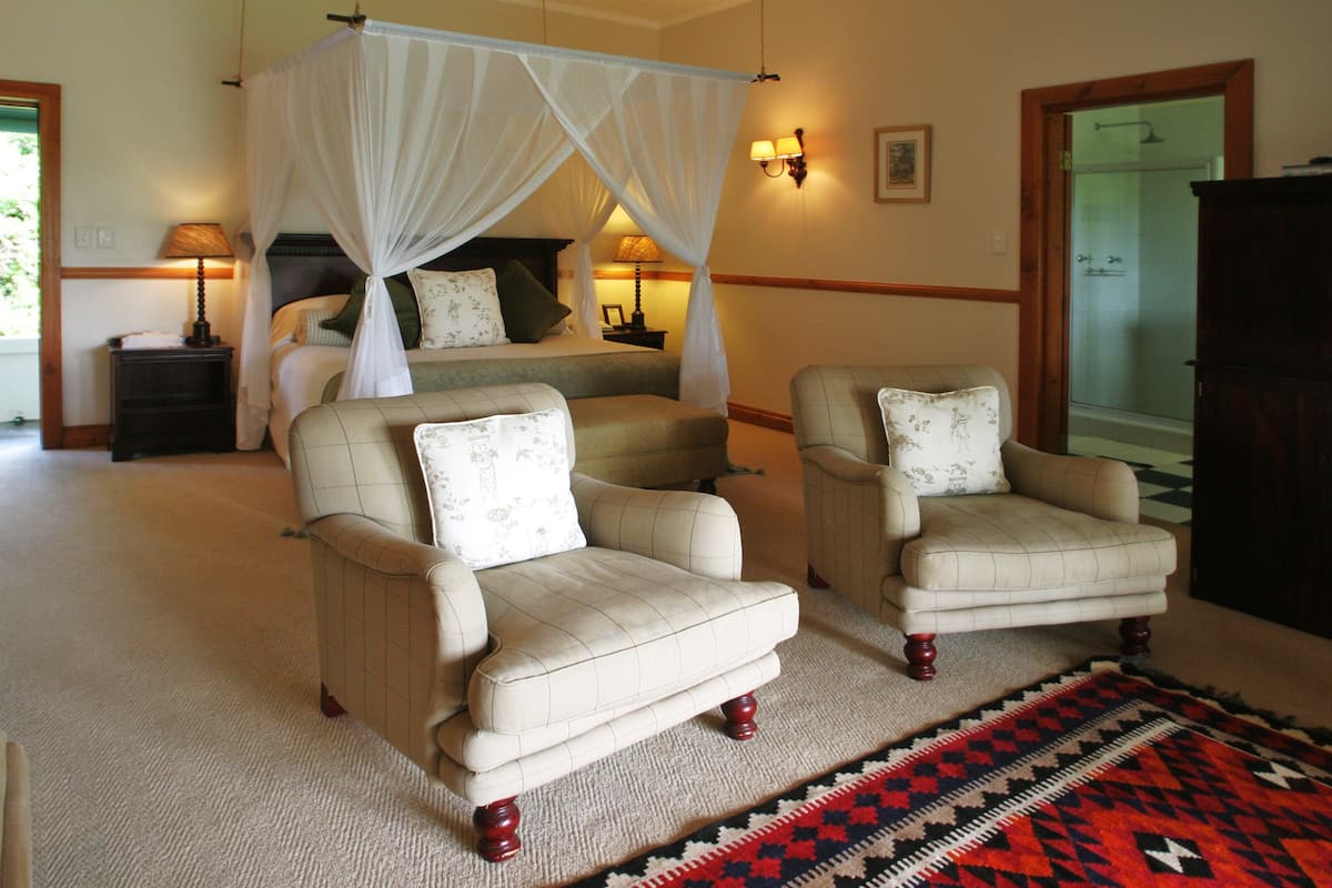 A bedroom with a canopy bed, two armchairs, a nightstand, and a patterned rug.