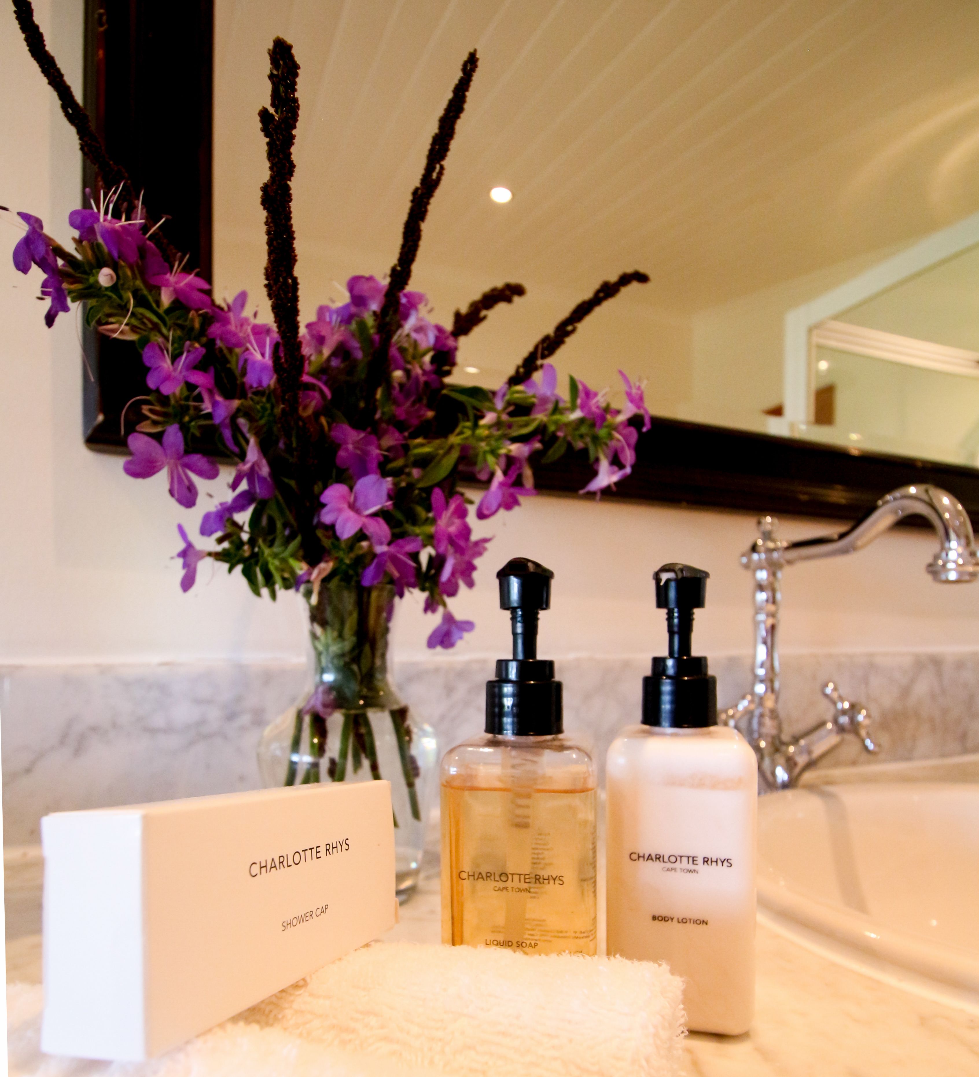 A bathroom with a white towel, a shower cap, and two bottles of Charlotte Rhys body lotion and liquid soap.