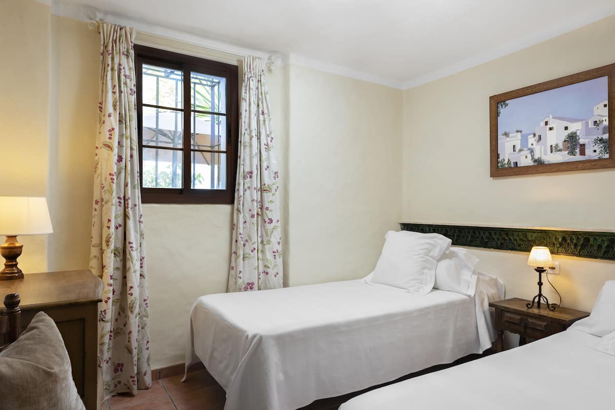 A hotel room with two beds, a window with curtains, a lamp on a bedside table, and a painting on the wall.