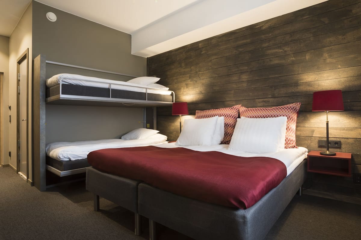 Ein Hotelzimmer mit Etagenbett, einem Einzelbett, einer roten Decke, zwei roten Lampen und einem Holz-Kopfteil.