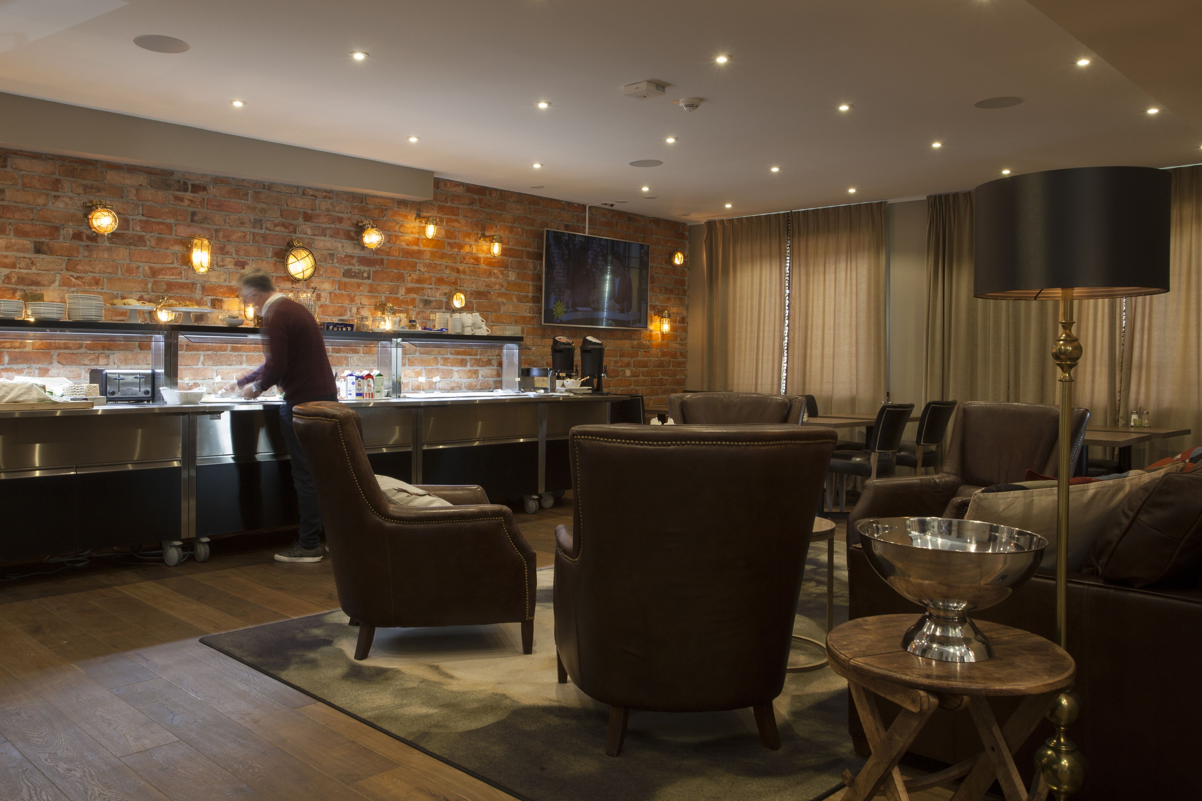 Una hall d'albergo moderna con una parete in mattoni, un'area bar, sedute in pelle e un'ampia lampada da terra.