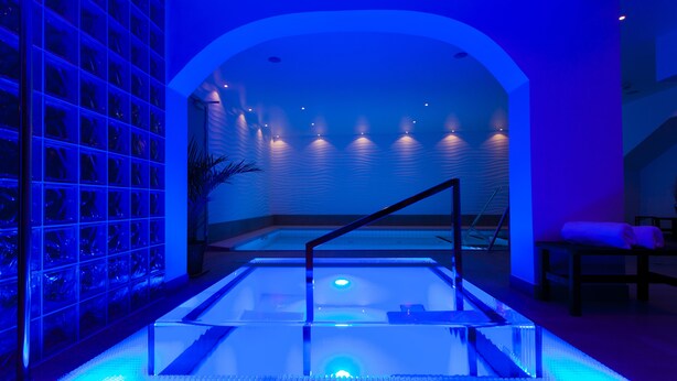 A modern spa area with a glass-enclosed hot tub, a seating area with a bench and cushions, and a wall with a grid of glass panels. - Hotell Nordic (Norrköping)