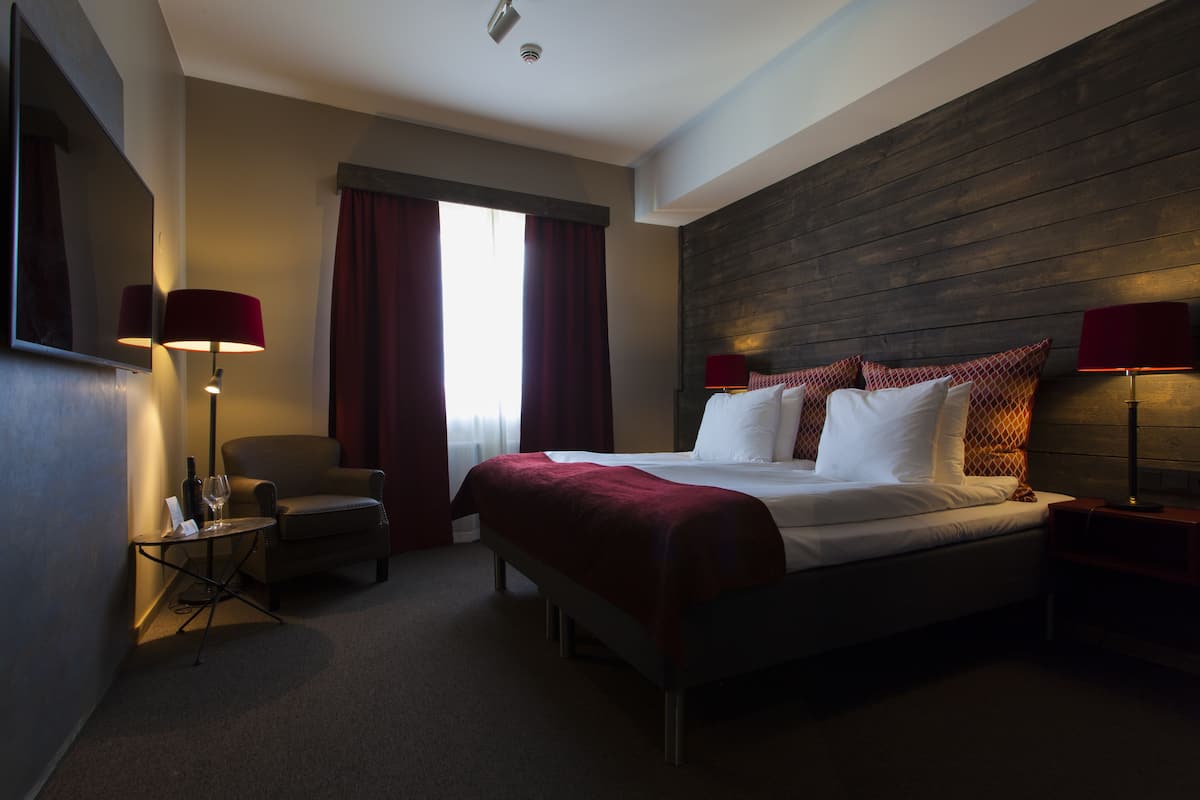 Ein Hotelzimmer mit einem großen Bett, zwei Nachttischen mit Lampen, einem Sessel und einem Fenster mit Vorhängen.