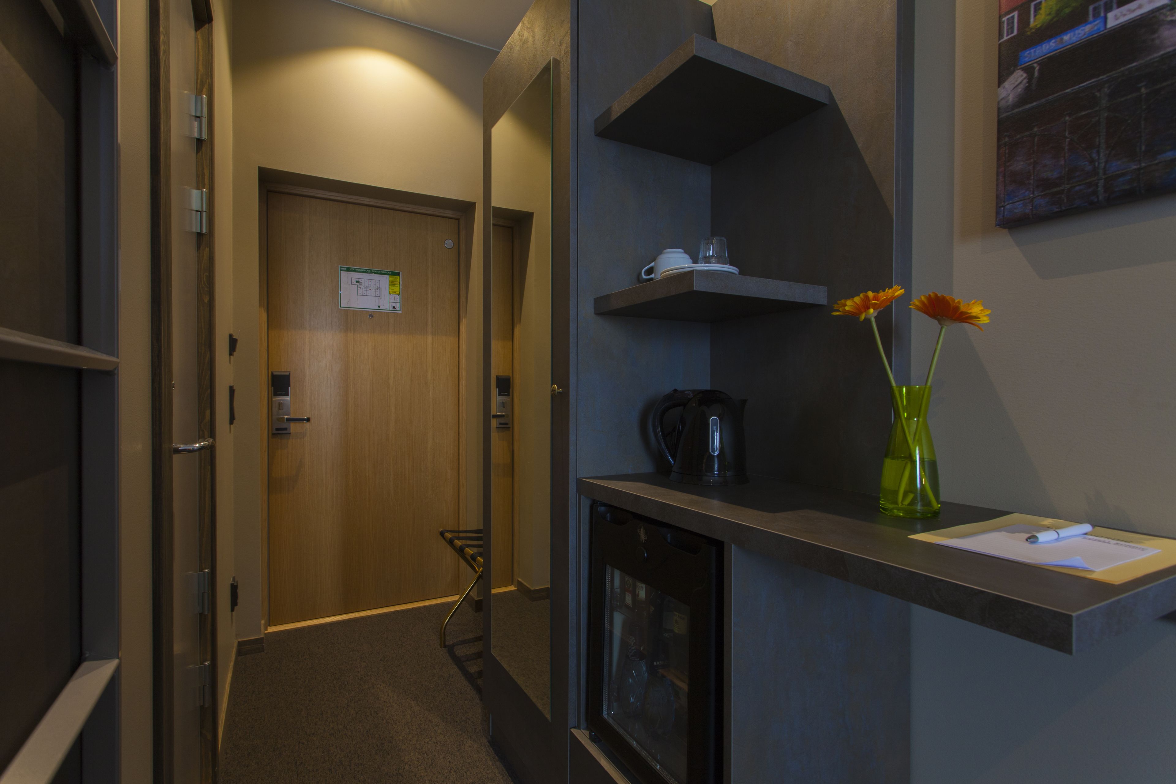 En hotellkorridor med en dörr av trä, ett litet pentry med en vattenkokare och en vas med blommor på en bänk.