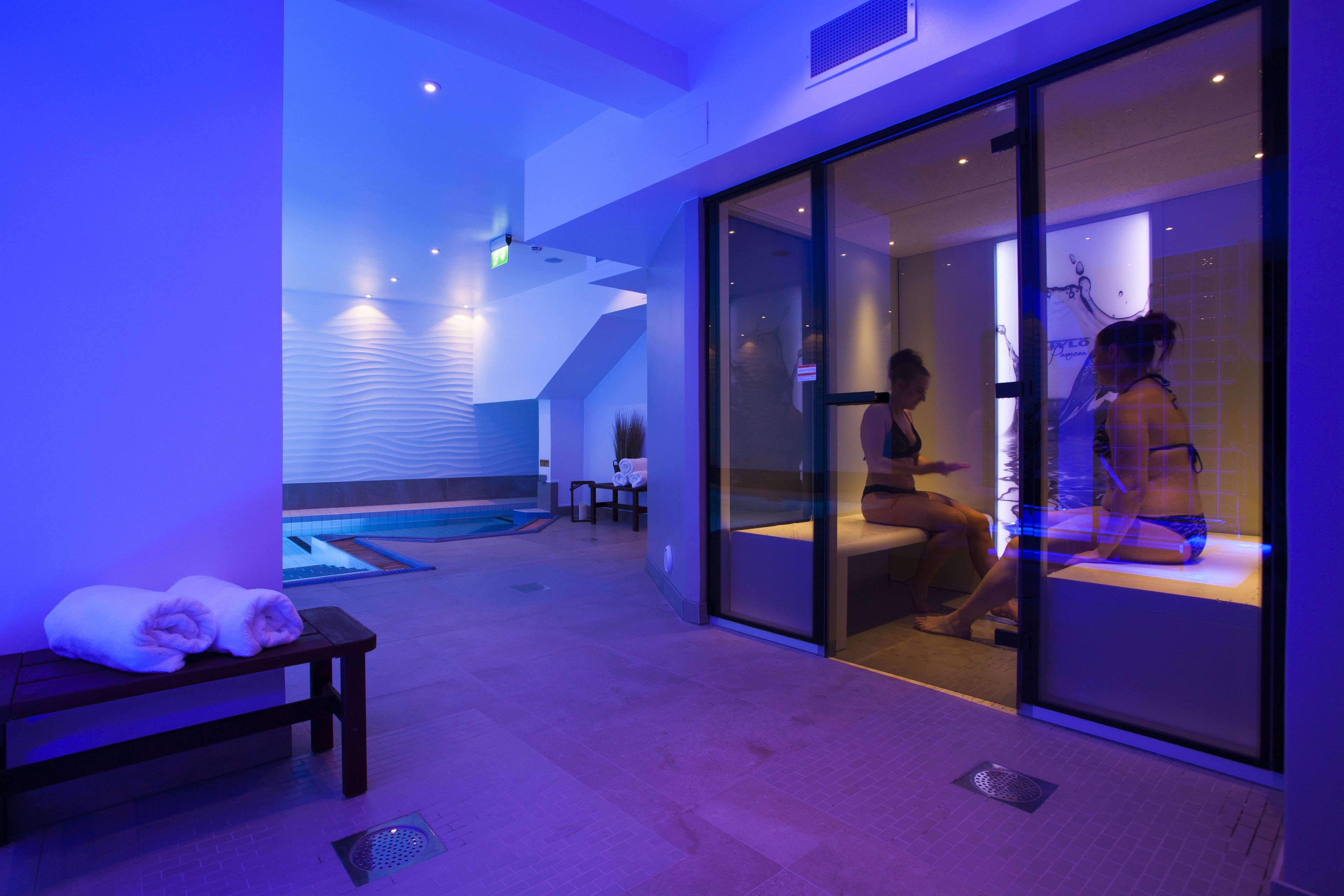 A spa area with a massage room, a bench with towels, and a pool in the background.