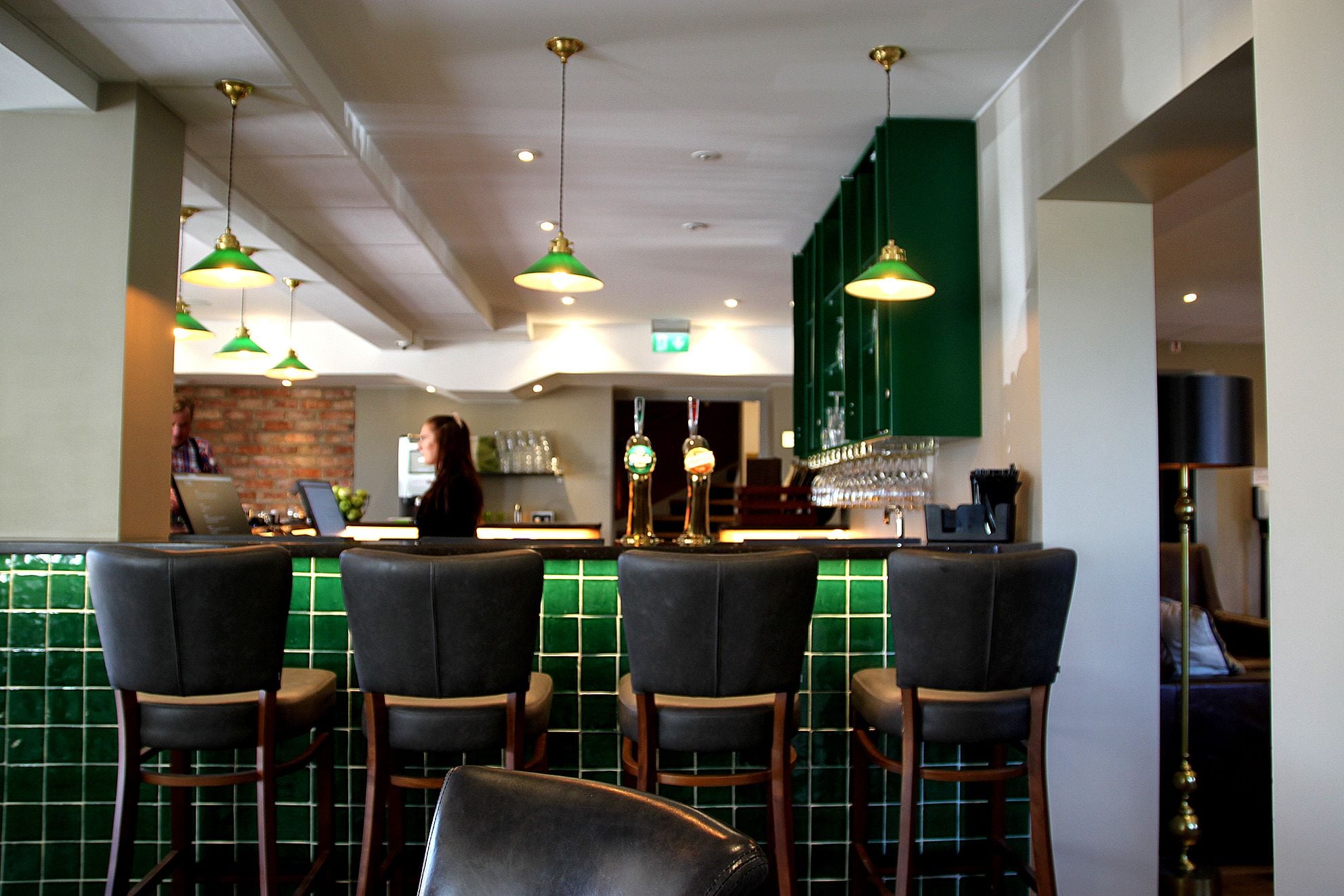 Zona de bar con paredes de azulejos verdes, taburetes de cuero negro e iluminación colgante verde.