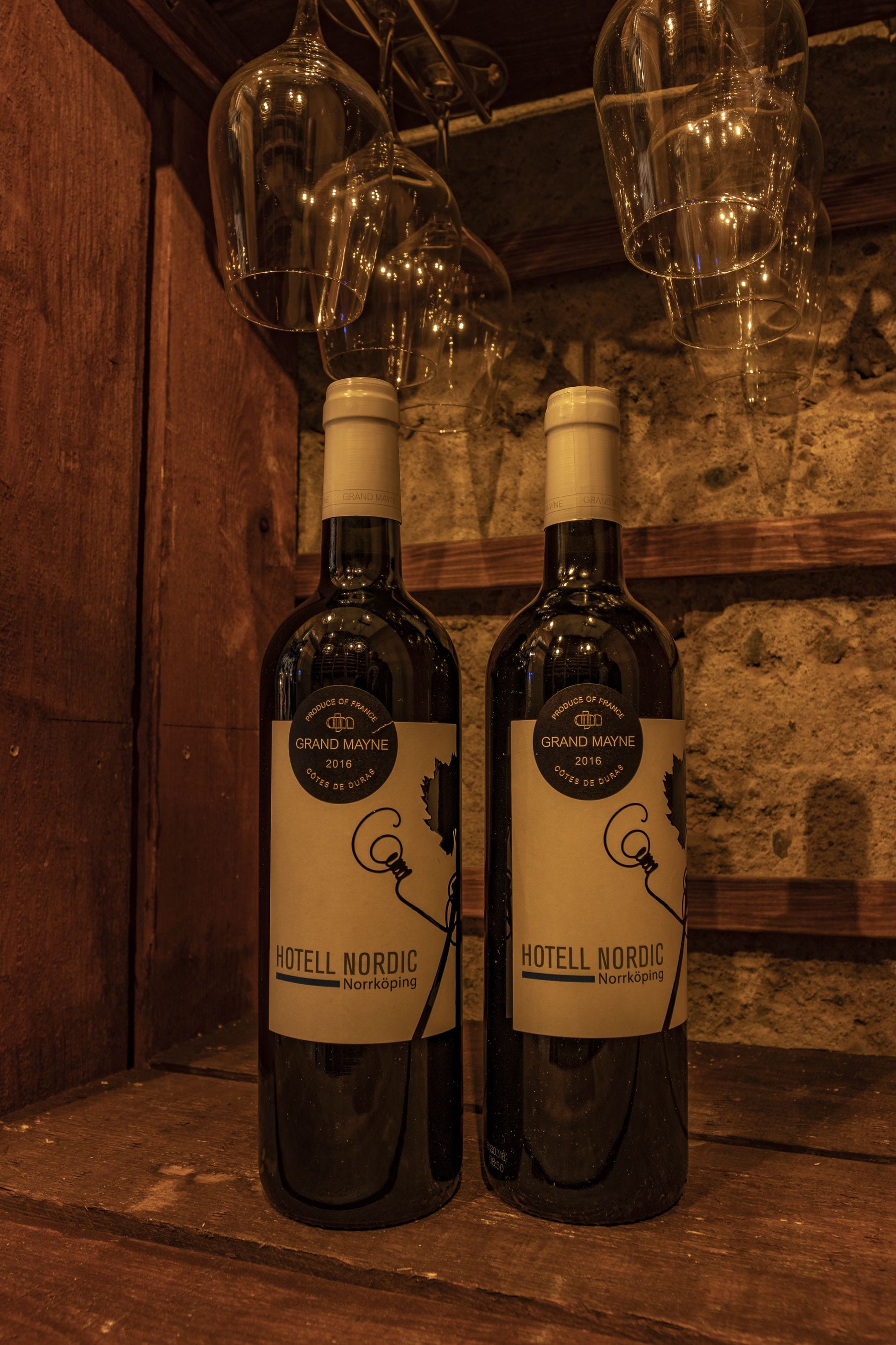 Två flaskor Grand Mayne-vin står på en trähylla, och i bakgrunden hänger vinglas.