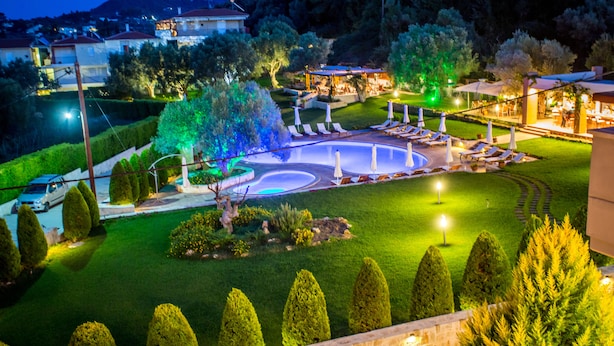 A well-lit outdoor pool area with lounge chairs and a fountain, surrounded by neatly trimmed hedges and trees. - Kassandra Village Resort (Kassandra)