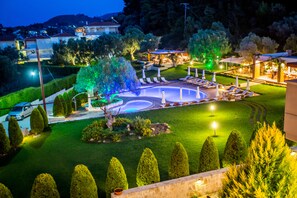 A well-lit outdoor pool area with lounge chairs and a fountain, surrounded by neatly trimmed hedges and trees. - Kassandra Village Resort (Kassandra)
