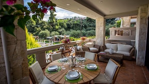A covered patio with a round wooden table set for dining, surrounded by wicker furniture and a stone fireplace. - Kassandra Village Resort (Kassandra)