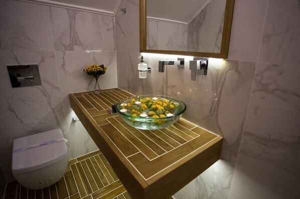 A bathroom with a marble wall, a wooden vanity, a glass bowl sink with yellow flowers, a toilet, and a mirror.