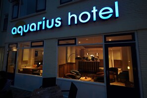 El exterior del Hotel Aquarius con señalización iluminada, amplias ventanas y un interior visible con asientos y una zona de bar.