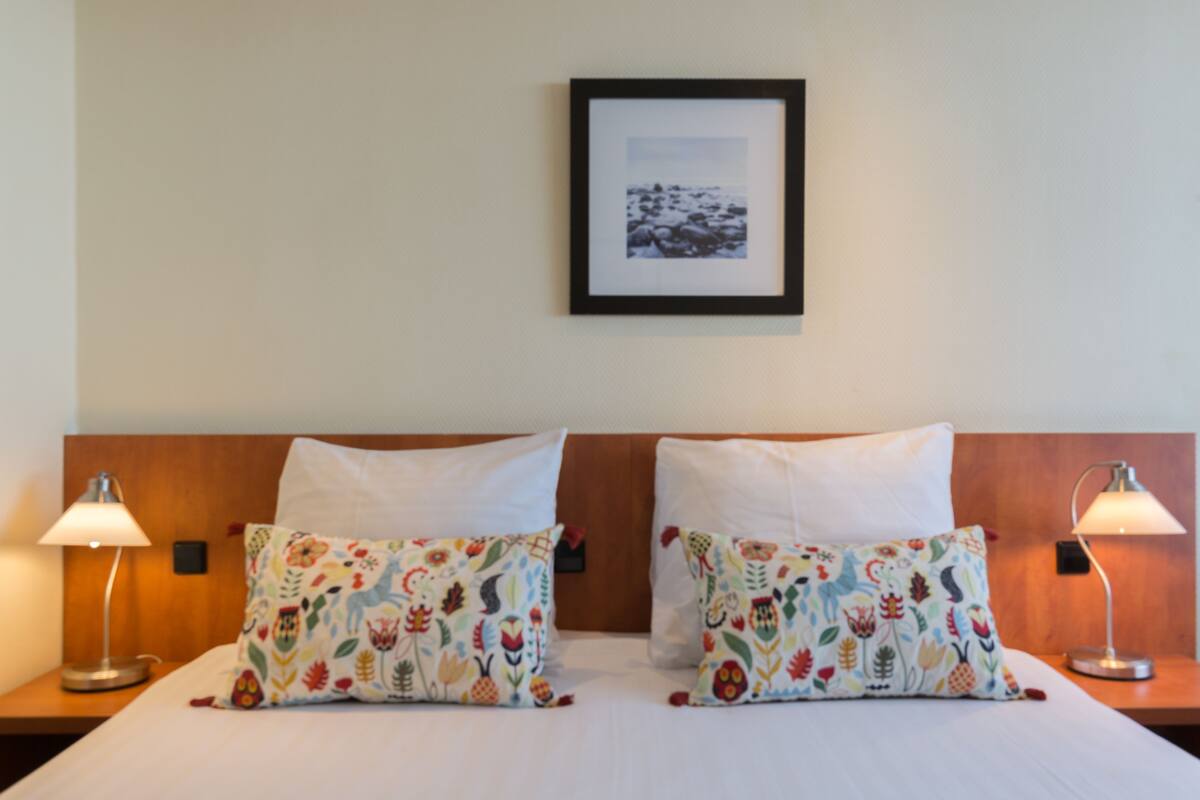 A hotel room with two beds, each with a white pillow and a colorful floral-patterned pillow. There is a framed picture on the wall above the beds, and two bedside lamps on either side of the room.