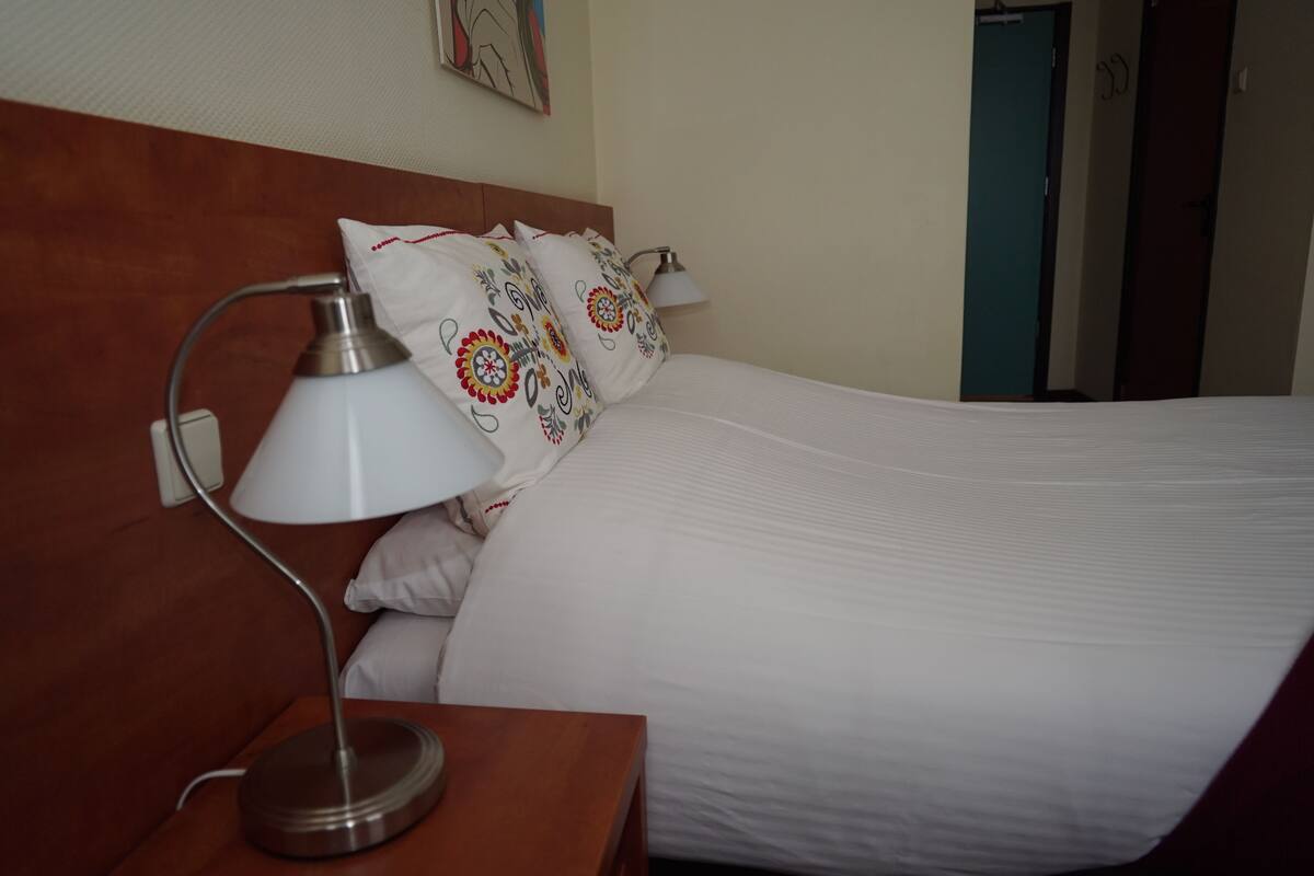 A neatly made bed with white linens and decorative pillows, a bedside lamp, and a wooden nightstand.