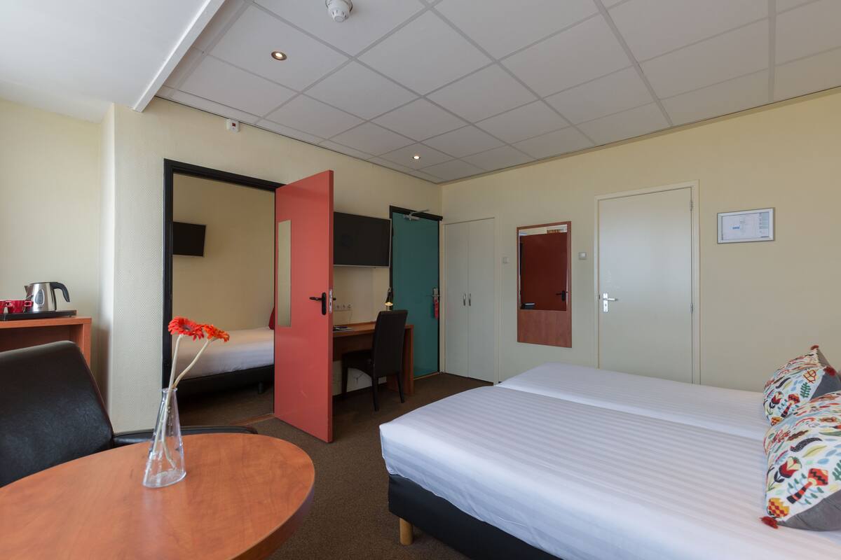 A hotel room with a bed, a desk, a chair, a small table with a vase, and a door leading to another room.