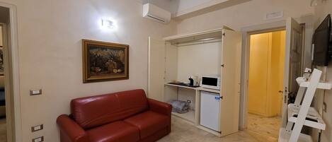 A room with a red leather sofa, a small kitchenette with a microwave, a refrigerator, and a television mounted on the wall.