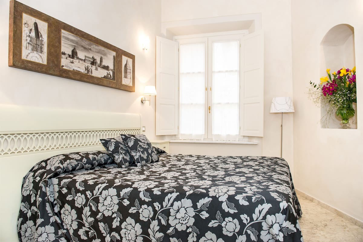 A bedroom with a bed, a window with shutters, a wall-mounted picture, and a bedside table with a lamp.