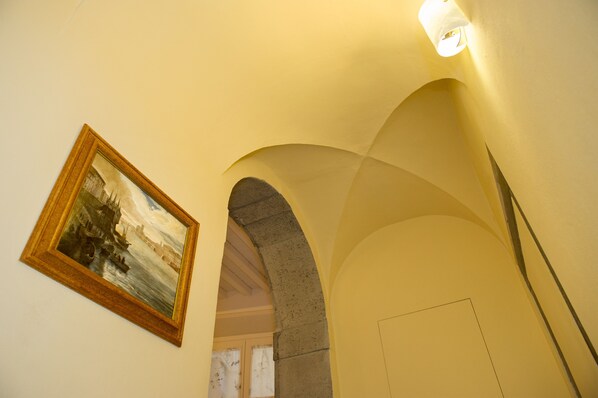 An interior corridor with a framed painting on the wall, an arched stone structure, and a light fixture. - Relais dei Mercanti B&B and Suites (Pisa)