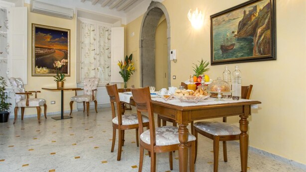 Een eetruimte met een marmeren vloer, houten stoelen, een tafel gedekt met fruit en brood, en schilderijen aan de wanden.