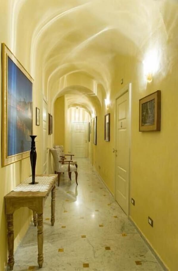 A hallway with a marble floor, framed artwork on the walls, and a wooden table with a vase. - Relais dei Mercanti B&B and Suites (Pisa)