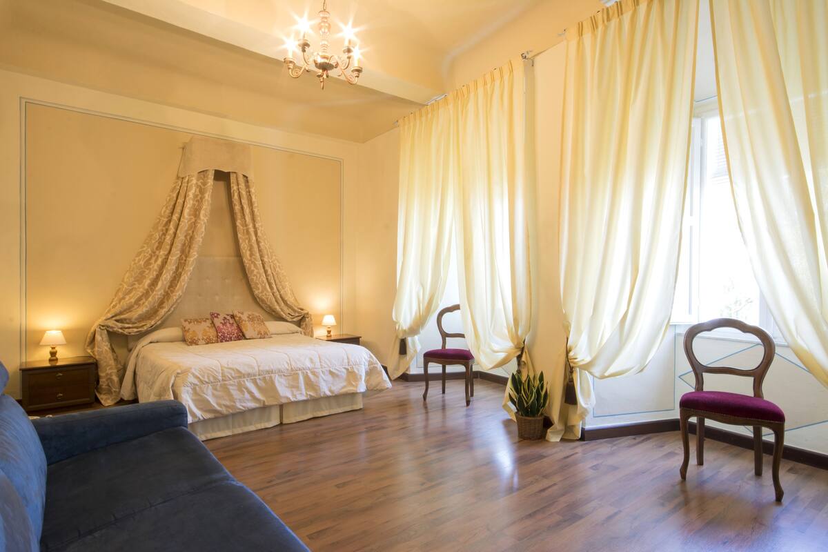 A bedroom with a canopy bed, a sitting area with a blue sofa, and a chair by the window.