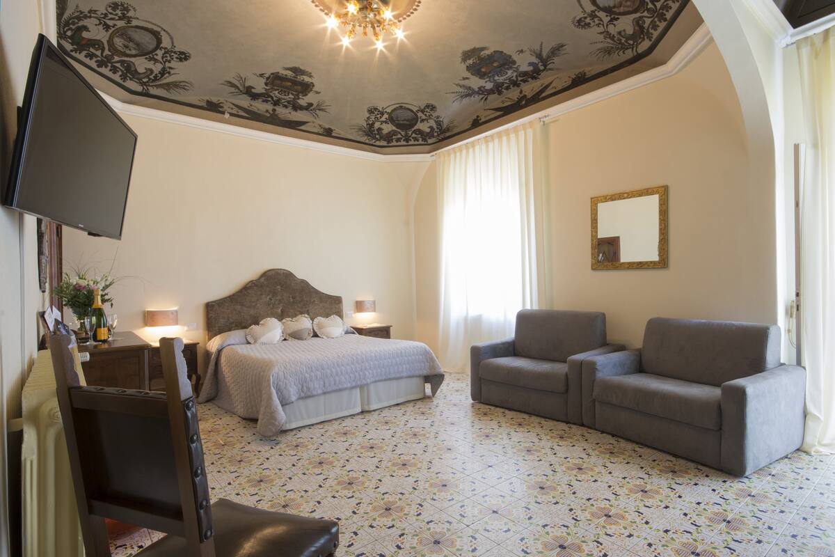 A hotel room with a bed, two armchairs, a TV, and a ceiling with floral patterns.