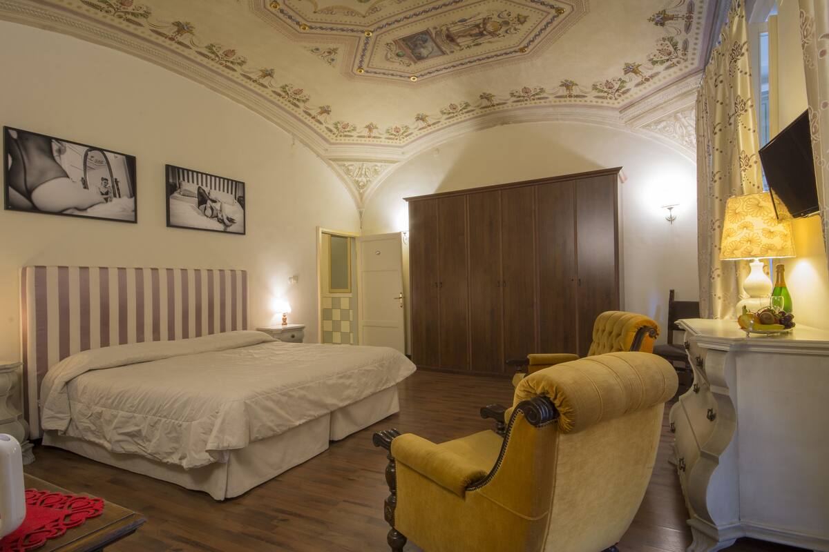 A hotel room with a large bed, a wooden wardrobe, a yellow armchair, and a flat-screen TV mounted on the wall.