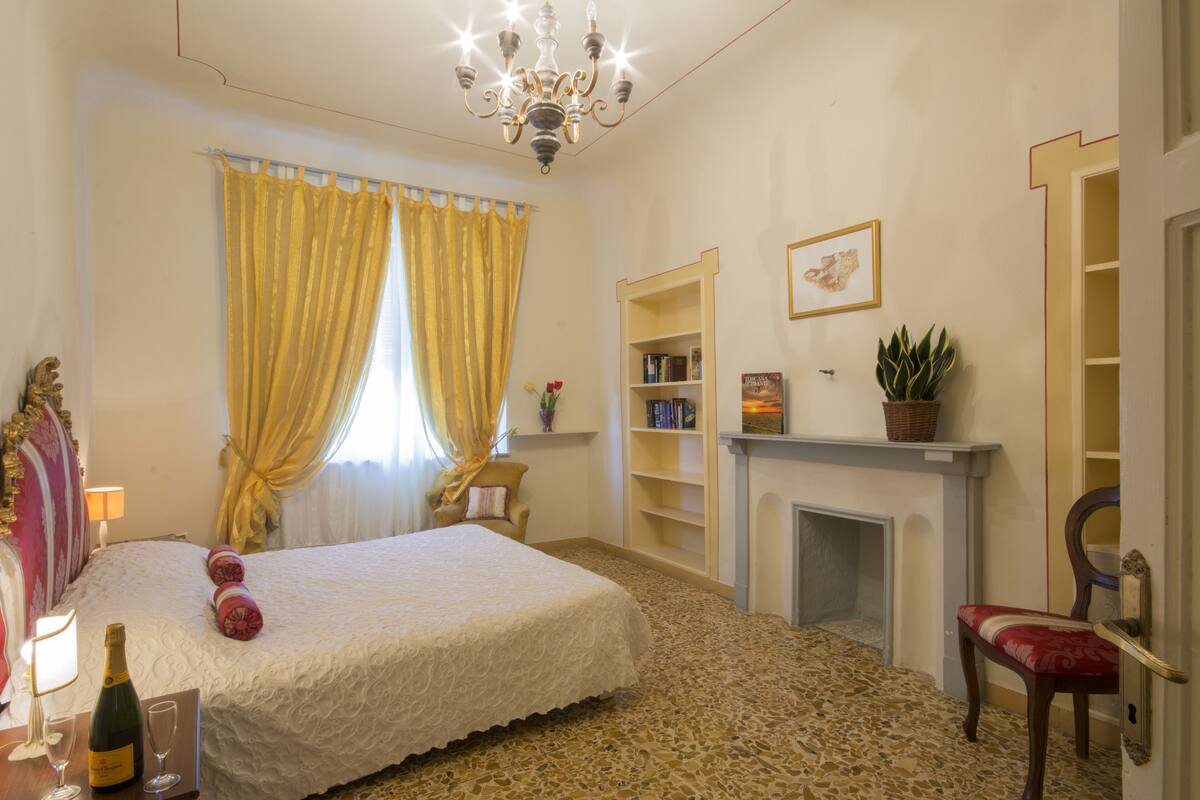 A hotel room with a bed, a chandelier, a fireplace, a bookshelf, and a chair.