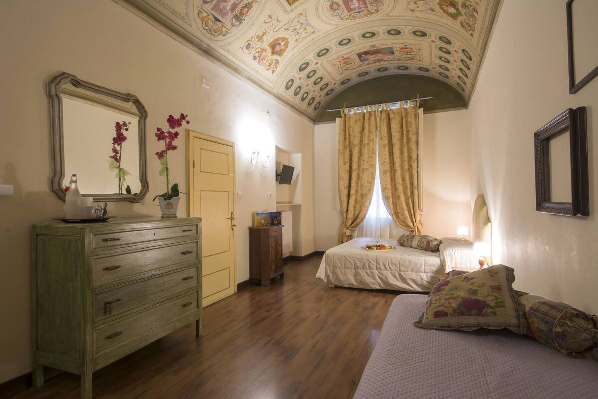 A bedroom with a wooden dresser, a bed with floral bedding, a ceiling with intricate artwork, and a door leading to another room.