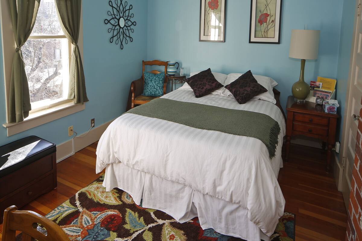 A bedroom with a bed, a chair, a nightstand, a lamp, and a window with curtains.