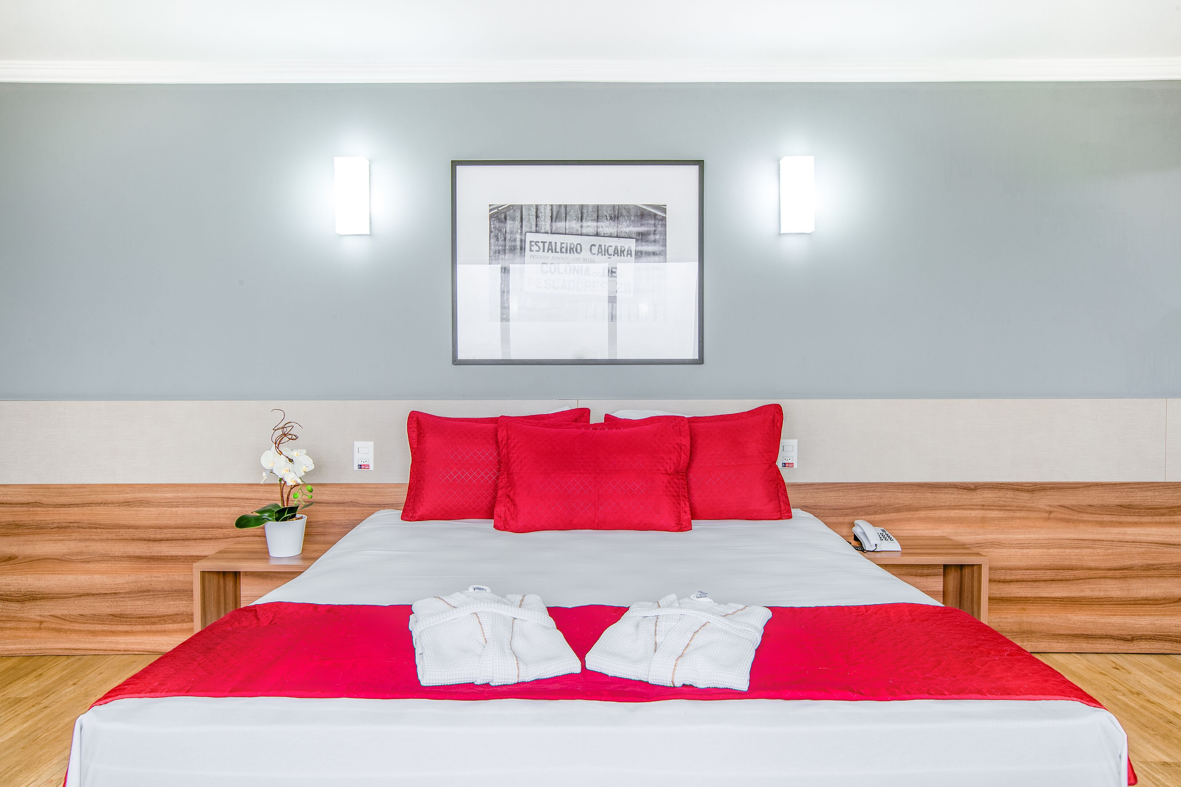 Un lit bien fait, avec une literie rouge et blanche, un tableau encadré accroché au mur et une petite plante en pot posée sur la table de chevet.