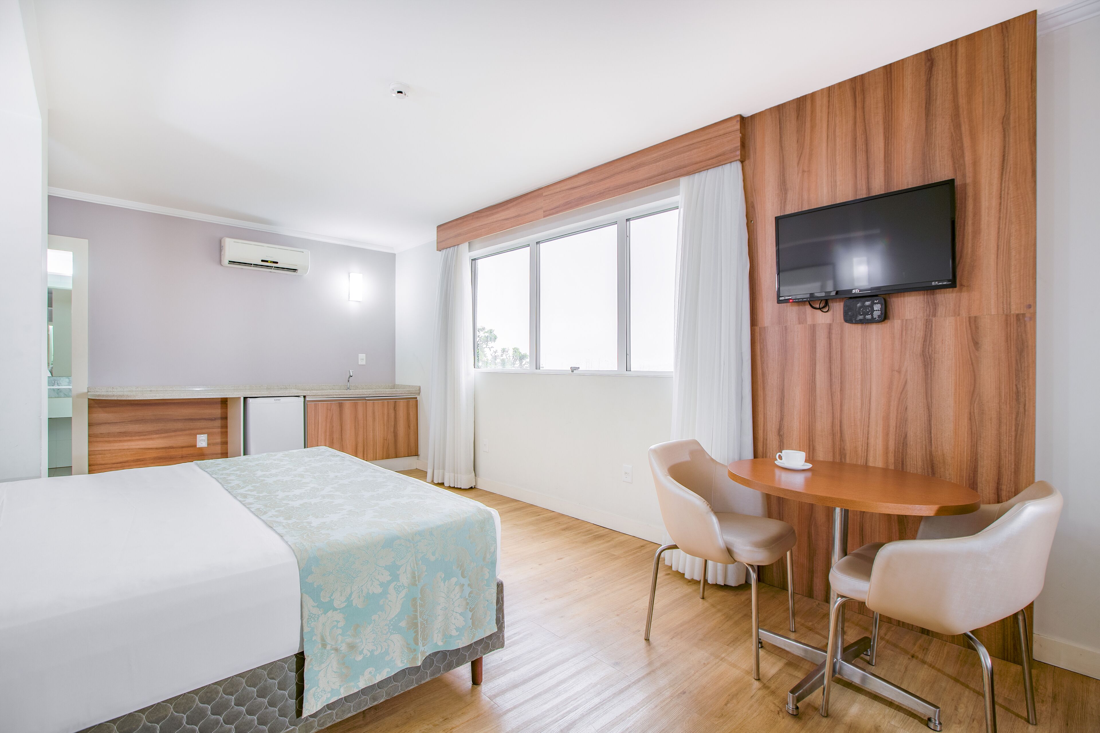 Quarto de hotel com cama, escrivaninha, cadeira, televisão e janela.