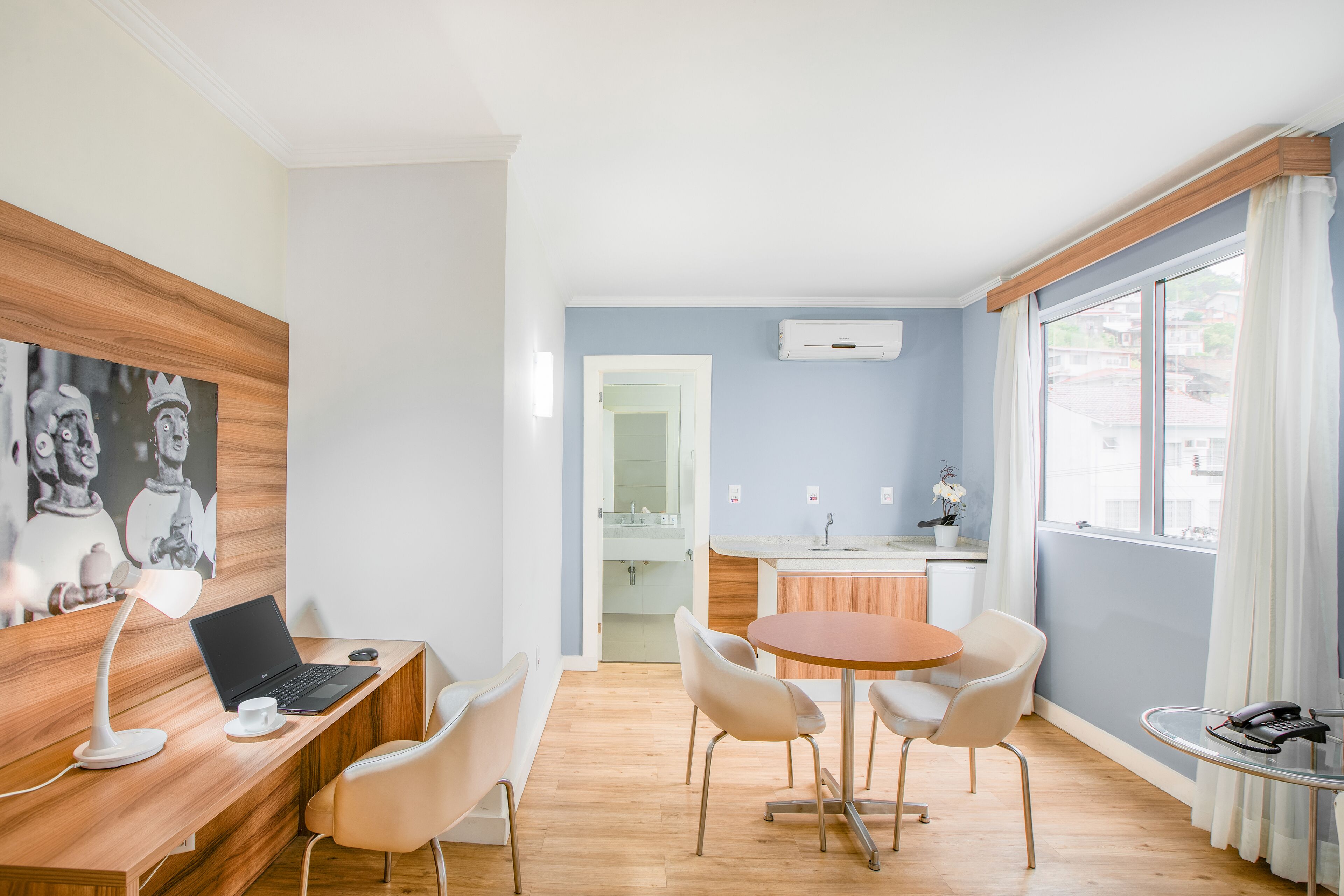 A modern room with a wooden desk, a laptop, a small round table with chairs, a window with curtains, and a kitchen area in the background.