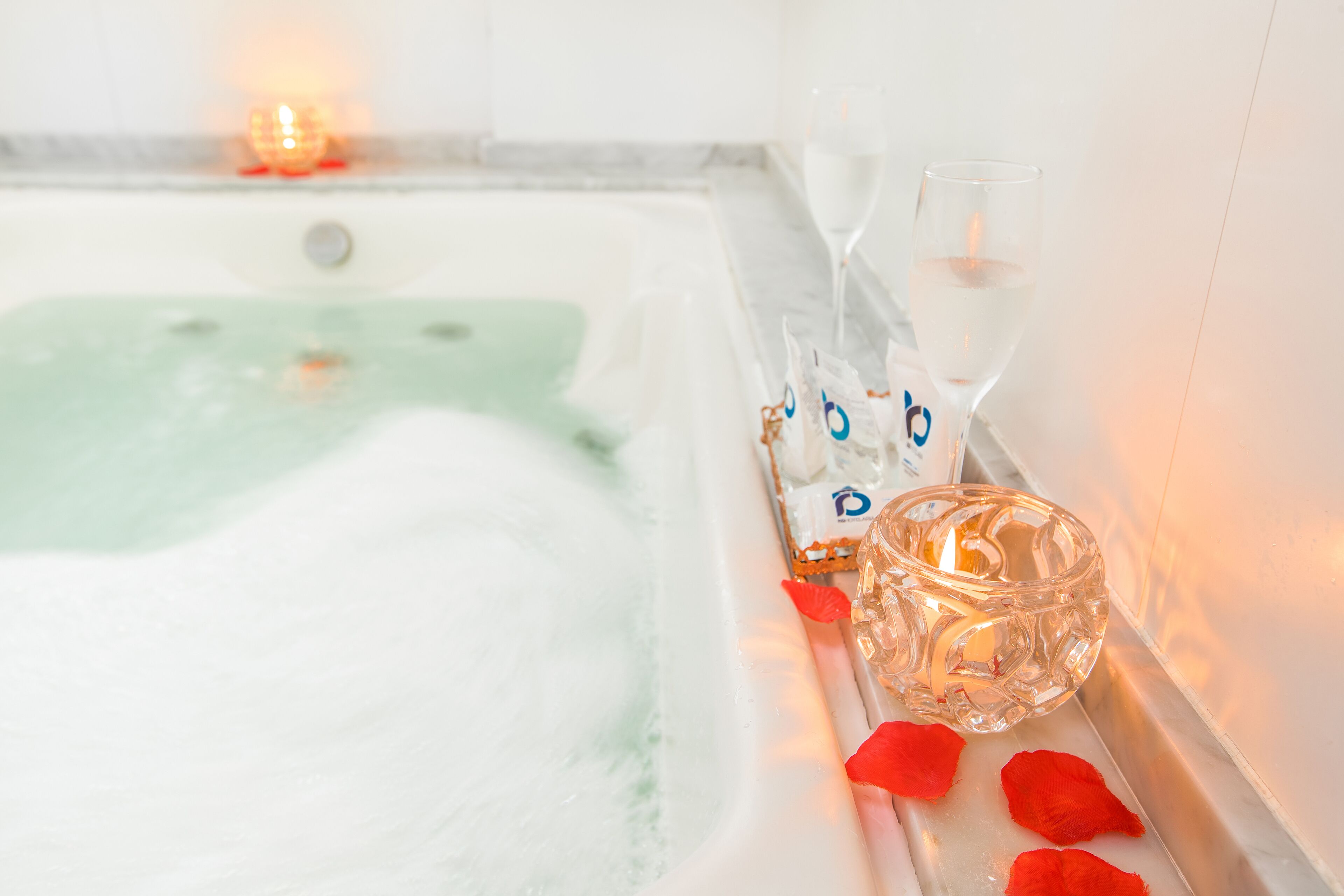 Una bañera llena de burbujas, un vaso de agua, una vela y pétalos de rosa.