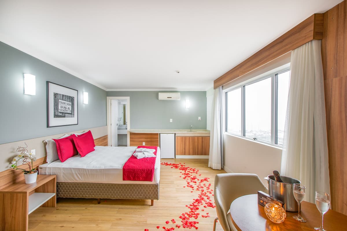 Ein Hotelzimmer mit Bett, Nachttisch, kleinem Schreibtisch, Stuhl, Fenster und einer Küchenzeile im Hintergrund.