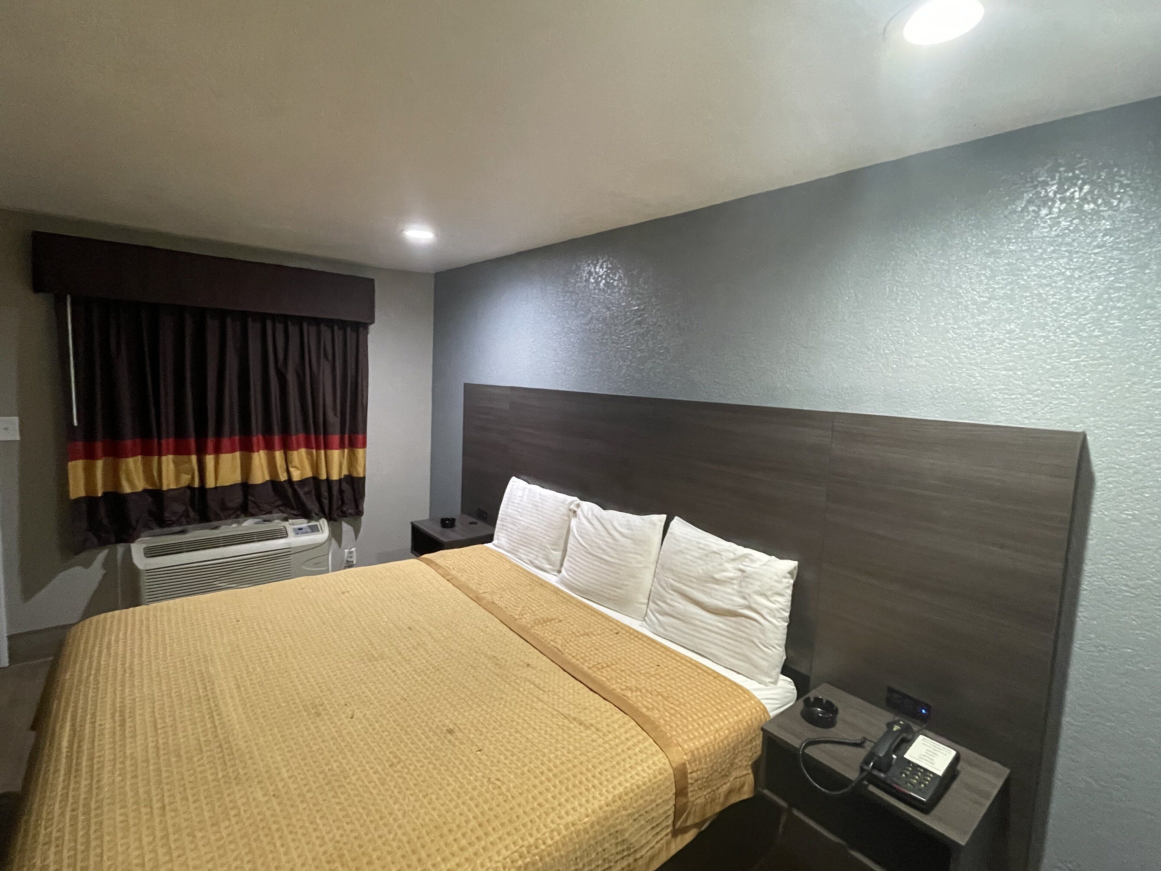 A hotel room with a large bed, a bedside table with a phone, a wall-mounted heater, and a window with curtains.