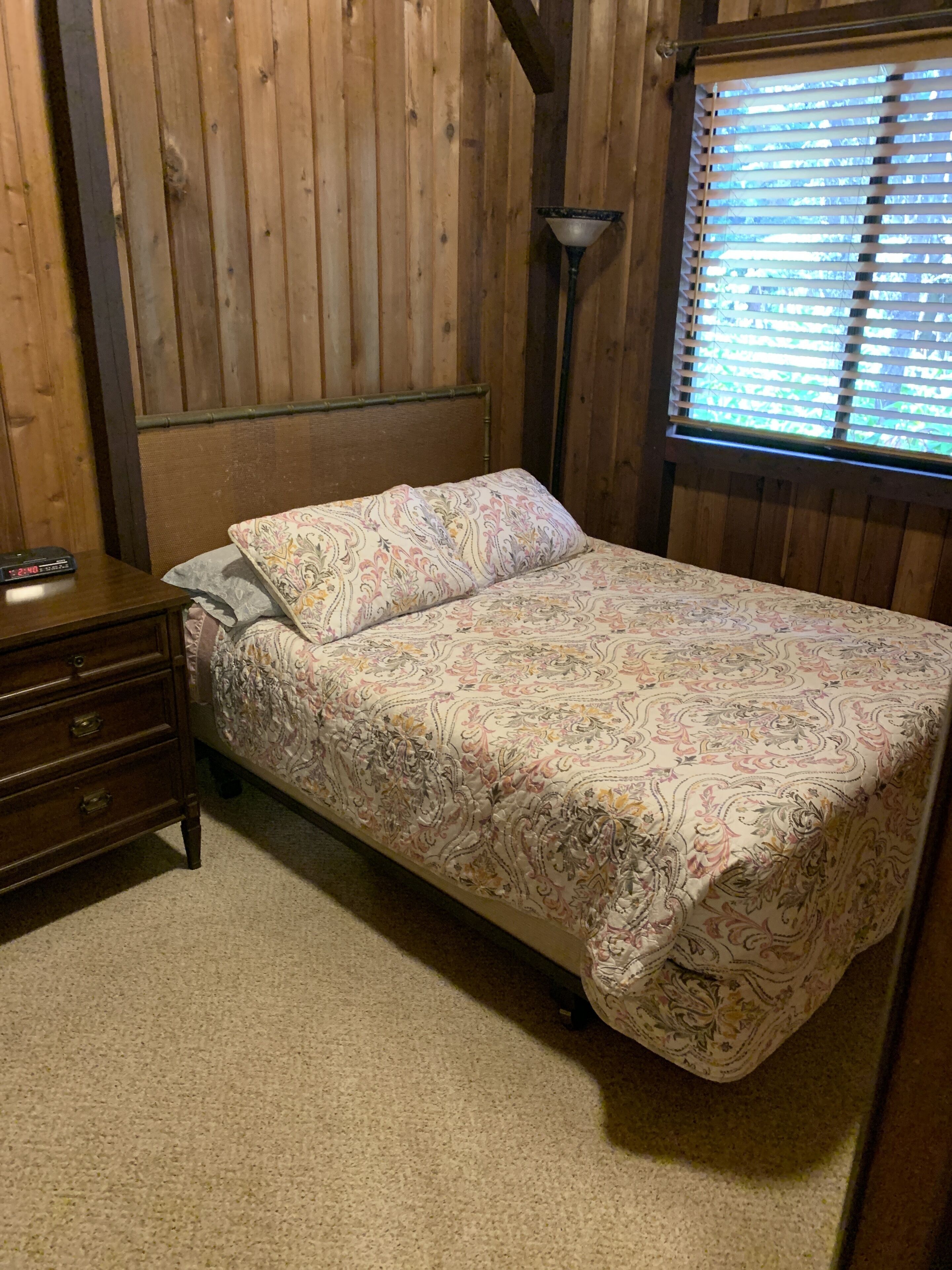 A bedroom with a wooden headboard, a bed with a floral bedspread, a nightstand with a lamp, and a window with blinds.