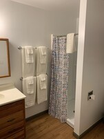 Un baño con tocador de madera, ducha con cortina estampada y varios toallas blancas colgadas en un perchero.